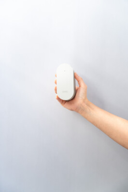 A hand holds a small, oval white electronic door lock by Simon Busse Studio for ABUS, on a plain light gray background.