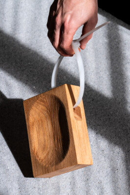 A hand holds the BO cutting board by Simon Busse Studio for Collect, with a zip tie handle, on a textured gray surface. 