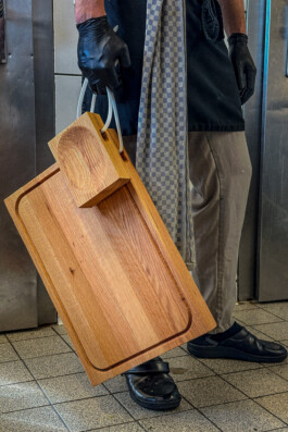 A person holds two wooden BO cutting boards by handles; design by Simon Busse Studio for Collect. Checkered towel on waist. 