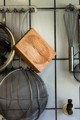 Metal kitchen utensils and a BO cutting board by Simon Busse Studio for Collect are neatly hung on hooks against white tiled wall. The cutting board hangs with a cable tie. 
