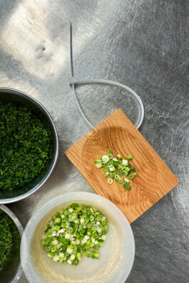 Chopped green onions on the BO cutting board by Simon Busse Studio for Collect, with bowls of herbs, on a metallic kitchen surface. 