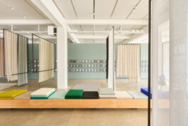 Simon Busse Studio portfolio: A modern gallery interior designed by industrial designer Simon Busse, featuring pastel green walls, three sheer hanging curtains that create a light, airy atmosphere, colourful bench cushions for flexible seating, and meticulously arranged framed artwork showcased in rows on the far wall.