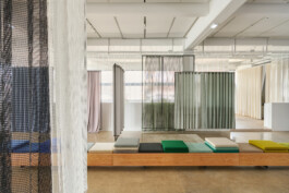 Simon Busse Studio portfolio image showing a modern interior with three layers of sheer curtains in different colours, a long wooden bench featuring multicoloured cushions designed with industrial precision, polished concrete flooring, and soft natural light highlighting the clean and functional aesthetic characteristic of Simon Busse Studio.