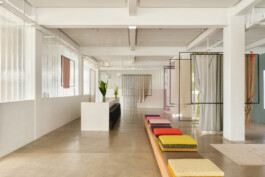 Simon Busse Studio industrial design showroom: a modern, minimalist space with polished concrete floors, crisp white walls, and suspended fabric panels framed architecturally. Three vibrant cushions rest on a long bench as inviting accents. At the rear, a counter displays vases and carefully placed plants, highlighting Simon Busse Studio’s signature blend of functional aesthetics and refined materiality.