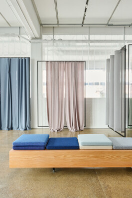 Portfolio image for Simon Busse Studio, industrial designer: A modern interior features a signature wooden bench by Simon Busse Studio, topped with three blue and grey cushions. Pastel curtains by the studio elegantly hang in front of expansive glass walls, dividing the luminous, industrial-inspired space with a refined design aesthetic.