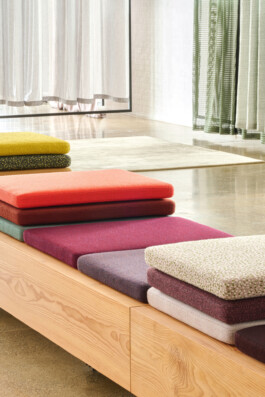 Three stacks of colourful seat cushions showcase Simon Busse Studio’s industrial design on a light wooden bench, set in a bright interior with polished floors, sheer curtains, and fabric panels in the background.