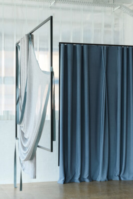 In an airy, industrial-style interior by Simon Busse Studio, three blue curtains hang closed on the right side, whilst a metal frame designed for functional aesthetics supports delicately draped light grey fabric with blue edges on the left.