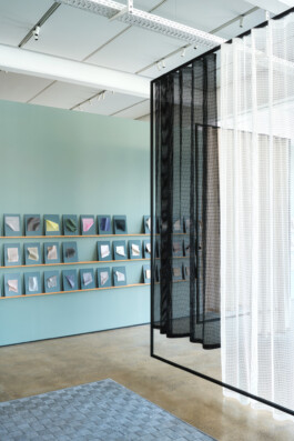 Simon Busse Studio portfolio: a modern gallery space with pastel blue walls features three small books arranged on precisely angled shelves, reflecting thoughtful industrial design. Mesh black and white curtains, reminiscent of innovative material choices, are paired with a sleek grey rug atop a polished concrete floor, highlighting the studio's signature blend of functionality and aesthetic clarity.