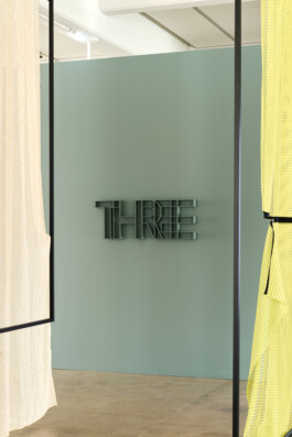 Simon Busse Studio portfolio: Industrial design interior with a pastel green wall and a bespoke black metal sign spelling THREE. The space features sheer beige and yellow curtains on either side, enhancing the minimalist atmosphere, with polished concrete flooring reflecting the studio’s refined aesthetic.