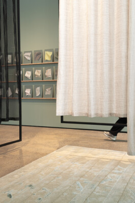 In Simon Busse Studio’s portfolio, a person in trainers walks behind a large cream-coloured curtain within a modern, minimalist room designed by industrial designer Simon Busse. Three fabric swatches are displayed on the wall, and a beige textured rug covers the floor, highlighting material selection and spatial composition.