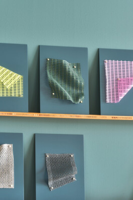 Simon Busse Studio presents an industrial design installation: three of four blue wall-mounted panels display textured and colourful fabric swatches—yellow, green, pink, and silver—pinned and draped in precise geometric patterns above a sleek, narrow wooden shelf.