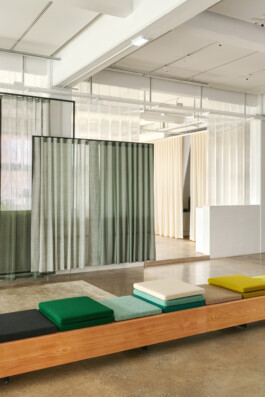 Simon Busse Studio portfolio: modern interior featuring a wooden bench with three vibrant cushions, set against a backdrop of sheer green and white curtains subtly dividing the space. A minimalist white reception desk appears in the background. The polished concrete floor enhances the industrial design aesthetic characteristic of Simon Busse Studio. 