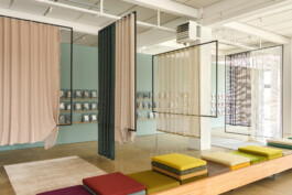 Simon Busse Studio portfolio project: Modern interior featuring three suspended fabric panels, pastel-toned walls lined with organised books or cards, and a bespoke long wooden bench fitted with vibrant seat cushions. The polished concrete floor and abundant natural light create an airy, contemporary setting characteristic of Simon Busse Studio’s industrial design aesthetic. 