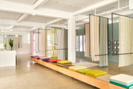 Simon Busse Studio: In a modern, bright showroom, three colourful seat cushions rest on a wooden bench designed with clean industrial lines. Neutral-toned hanging fabric panels by Simon Busse Studio divide the space, showcasing innovative spatial solutions and contemporary material combinations for this industrial designer’s portfolio. 